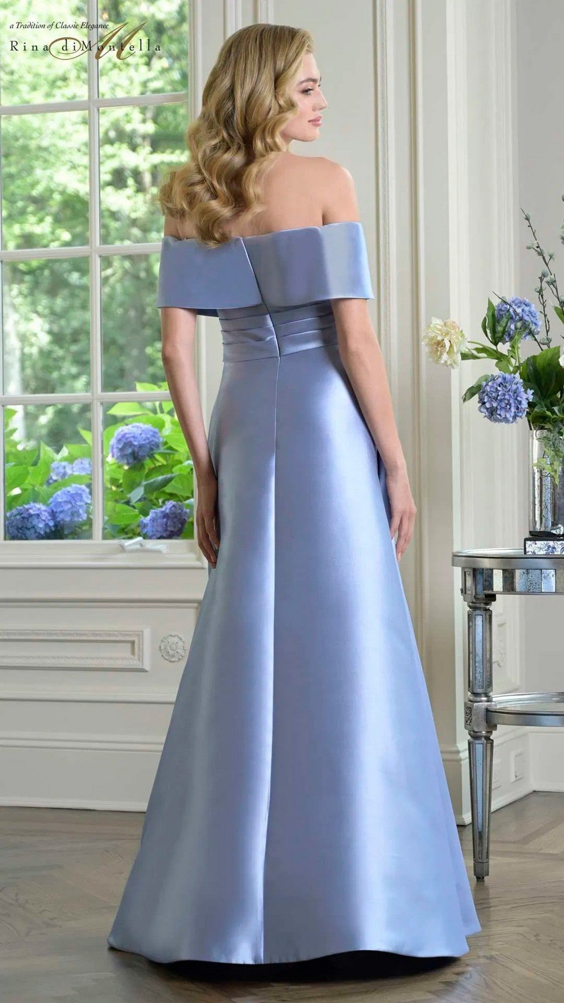 A woman indoors, near a window and flowers, faces away from the camera wearing the light blue Rina Di Montella RD3429 long formal off-the-shoulder dress with a flared skirt.